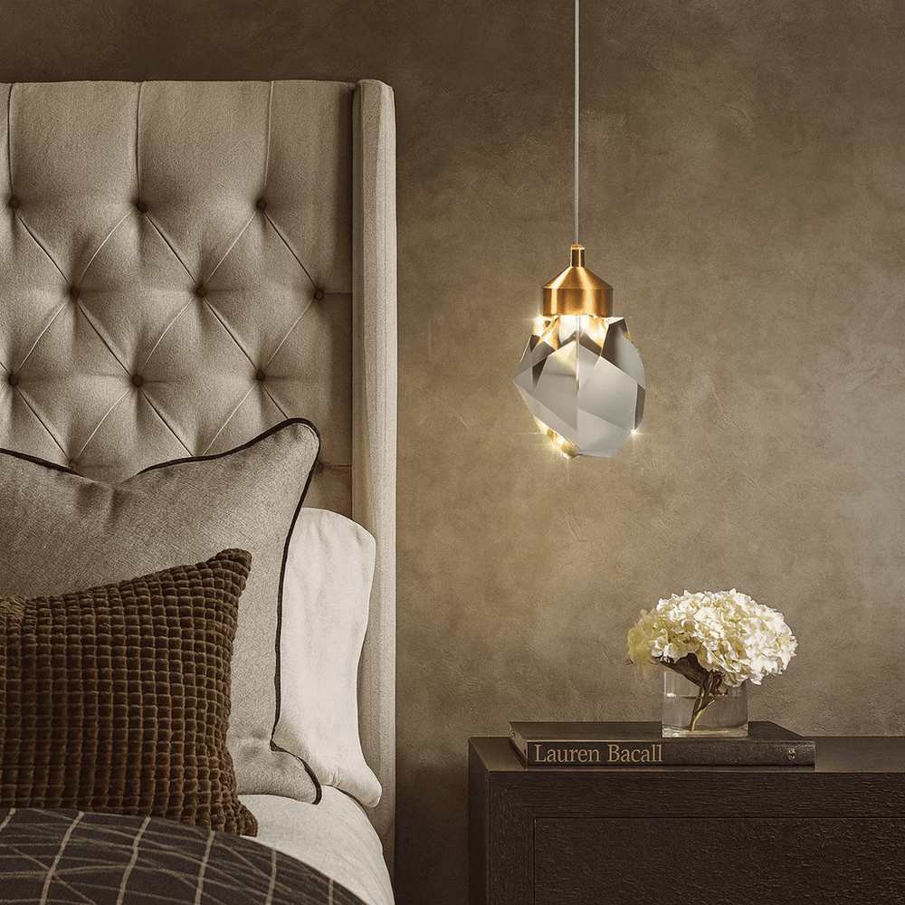 A modern bedroom features a tufted headboard, brown textured pillows, the Morsale Valenza Grand Crystal Pendant for luxurious lighting, a side table with a white flower arrangement, and the book “Lauren Bacall.”.