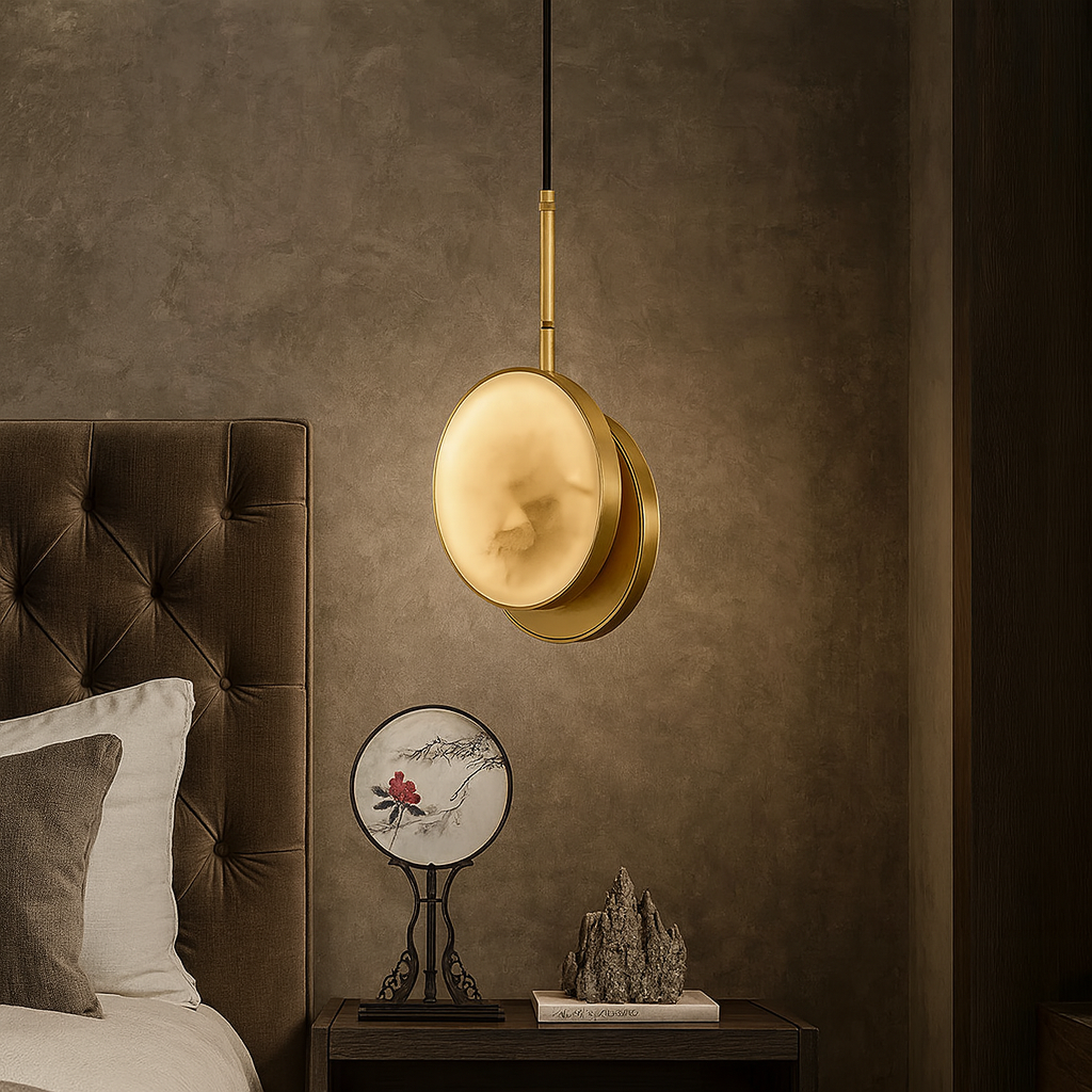 A Morsale.com Aeris Marble Pendant Light in brushed brass hangs above a bedside table with a plate, book, and rock sculpture next to a tufted brown headboard and beige pillow in this softly lit, elegant bedroom.