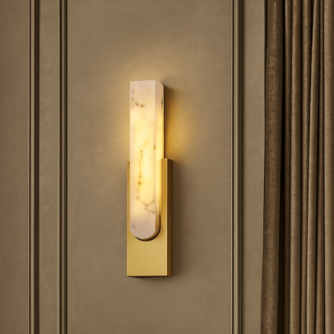 The Wexford Marble Wall Sconce from Morsale.com features a rectangular Spanish marble shade and gold base, casting a warm LED glow on a taupe paneled wall beside brown curtains.