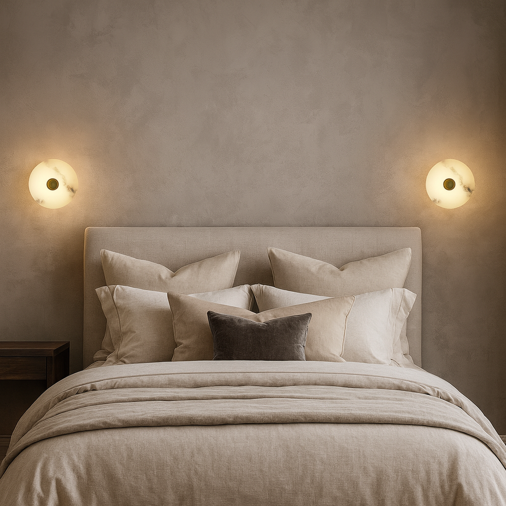 A neatly made bed with beige bedding and multiple pillows sits against a taupe wall, flanked by two Hastings Marble Wall Sconces from Morsale.com, providing warm, cozy lighting.
