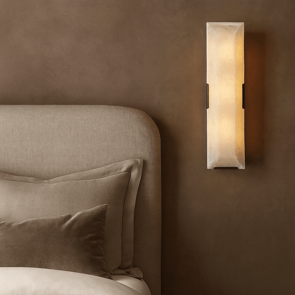 The Brookhill Calcite Wall Sconce by Morsale.com casts warm light above a beige upholstered headboard and pillows, set against a brown textured wall—an inviting addition of modern lighting.