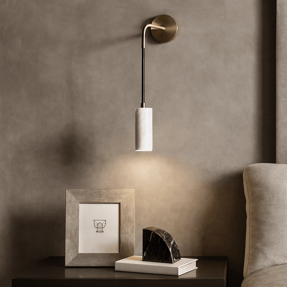The Biltmore Marble Pendant Wall Sconce by Morsale, featuring a cylindrical white marble shade, hangs above a dark bedside table with a framed minimalist drawing and marble sculpture, beside a beige upholstered bed.