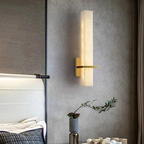 A modern bedroom wall features a vertical rectangular Morsale.com Natural Marble Indoor Sconce with a soft yellow-white LED indoor lighting and metallic accent. Below it, a small grey pot with greenery sits on a table. Part of a bed with a white headboard and grey bedding is visible on the left.