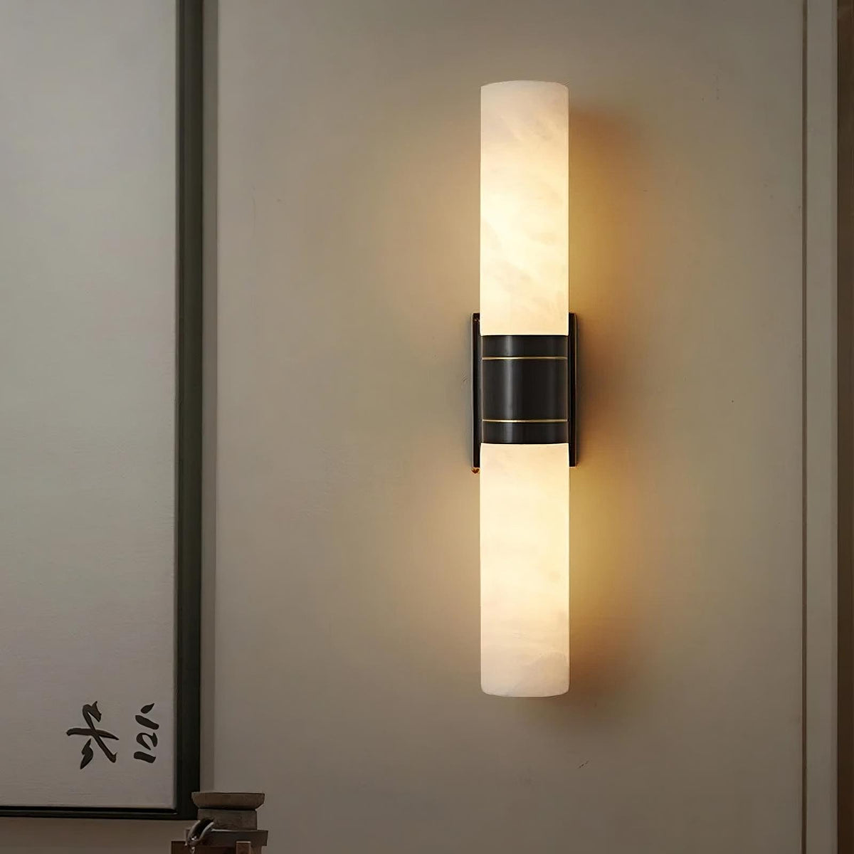 A Morsale.com Natural Marble Indoor Wall Sconce Light with two cylindrical white shades is mounted vertically on a beige wall. The light fixture, featuring a brass light source and black metal bracket, creates a cozy ambiance with its soft warm glow. Asian-style calligraphy is visible nearby, adding to the unique texture of the decor.