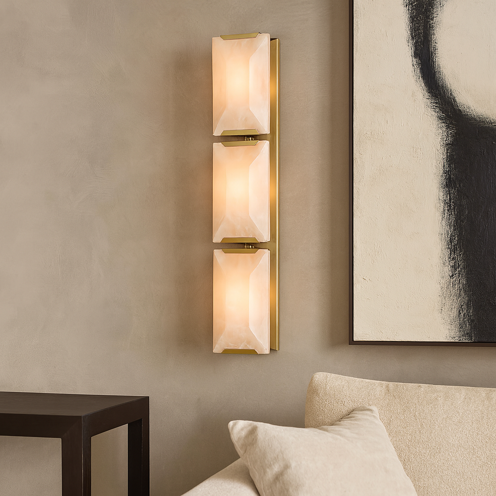 The Brookhill Calcite Crystal Sconce by Morsale.com features three rectangular frosted glass lights on a brass fixture, shown mounted on a beige wall beside a cream sofa and abstract black-and-white artwork, near a dark wood table.