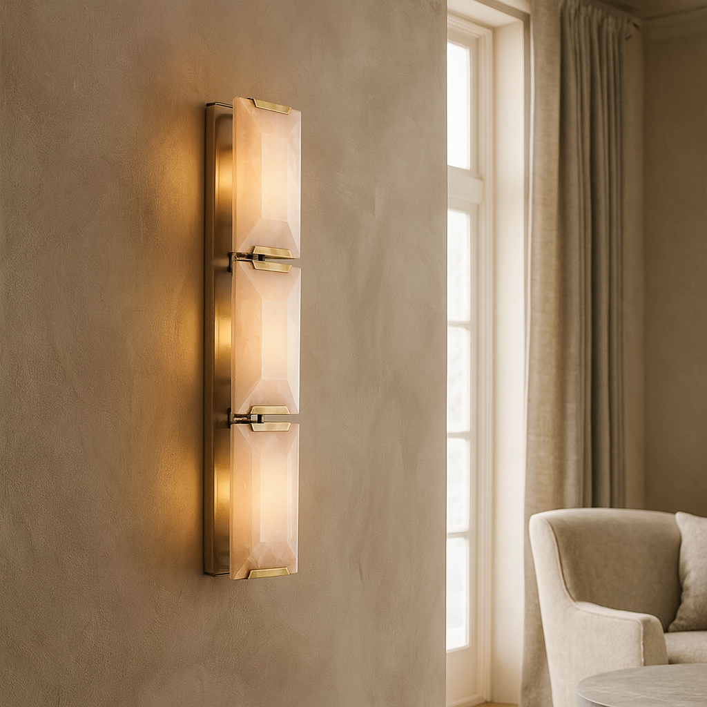 The Brookhill Calcite Crystal Sconce by Morsale.com features three rectangular crystal panels and brass accents, casting a warm glow on a beige textured wall beside a window with long curtains and a light upholstered chair.