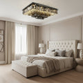 A modern bedroom featuring a tufted beige bed, neutral bedding, bedside tables with lamps, large curtained window, abstract wall art, and the eye-catching smoke grey Gio Crystal Flush Mount Chandelier by Morsale.com on the ceiling.