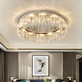 A modern living room features a large, circular Gio Flush Mount Crystal Ceiling Light from Morsale.com hanging from the ceiling. The light casts a warm glow with E14 LED Bulbs, reflecting off the crystals. Below, a framed abstract painting hangs on the wall above a green sofa adorned with cushions and a small floral arrangement.