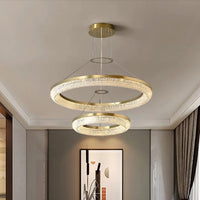 The Morsale.com Bergamo Crystal Chandelier elegantly illuminates a minimalist room with its energy-efficient design, featuring dual gold and clear crystal rings at varied heights. A contemporary artwork adorns the creamy walls, while dark curtains add a touch of elegance to the windows.