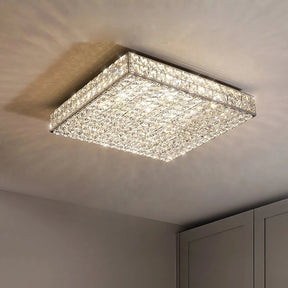 A square, Bacci Clear Crystal Light Fixture from Morsale.com is mounted on the ceiling, emitting bright light. The ceiling and part of a wall-mounted cabinet with gray doors are visible in the background. The light creates a reflective pattern on the ceiling.