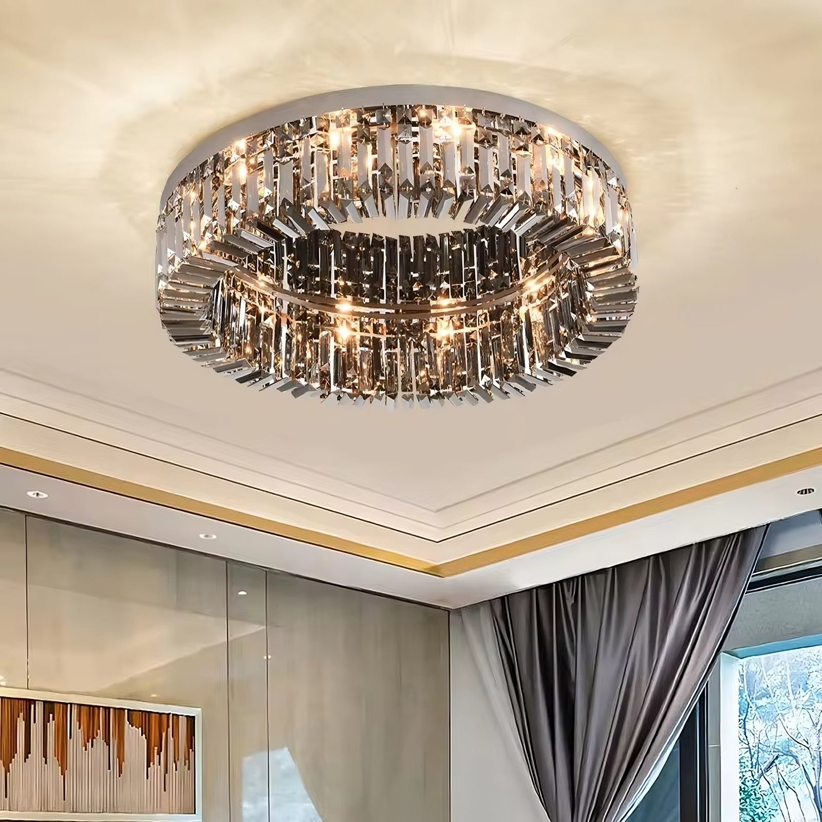 A Specchio Smoke Grey Crystal Ceiling Light from Morsale.com with cascading metallic elements hangs from the ceiling of a well-lit room. The ceiling features recessed lighting, and there's a large window with gray curtains to the right and a piece of abstract art on the wall to the left.