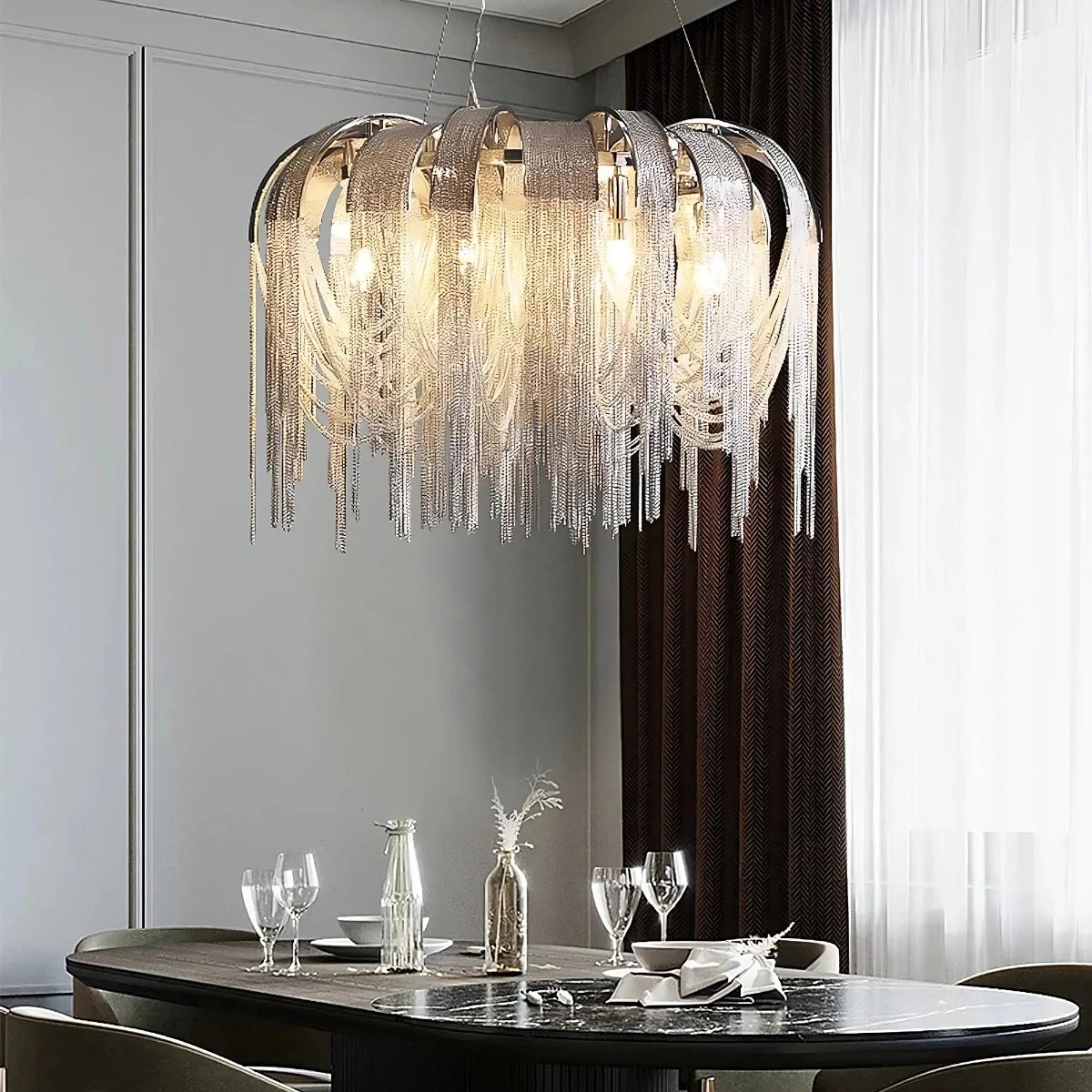 A modern dining room showcasing a Morsale.com Cadena Contemporary Chandelier, featuring adjustable hanging wire and cascading metallic strands with LED bulbs illuminating a dark oval table set with elegant glassware and minimalistic décor. The backdrop is adorned with floor-to-ceiling curtains and a light grey wall.