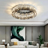A modern living room features a stunning Giano Crystal Ceiling Light Fixture by Morsale.com, its crystal-like drops hanging elegantly from a circular frame. The room has a white sofa adorned with yellow and green cushions, a white coffee table with a flower vase, and a green abstract painting on the wall.