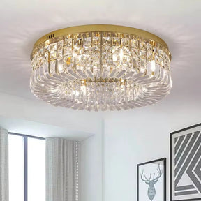 A luxurious Gio Crystal Ceiling Light from Morsale.com, featuring an opulent gold frame, is installed on a white ceiling in a modern interior. The room, illuminated by LED bulbs, showcases large windows with sheer curtains and geometric artwork of a deer on the wall, enhancing its contemporary decor.