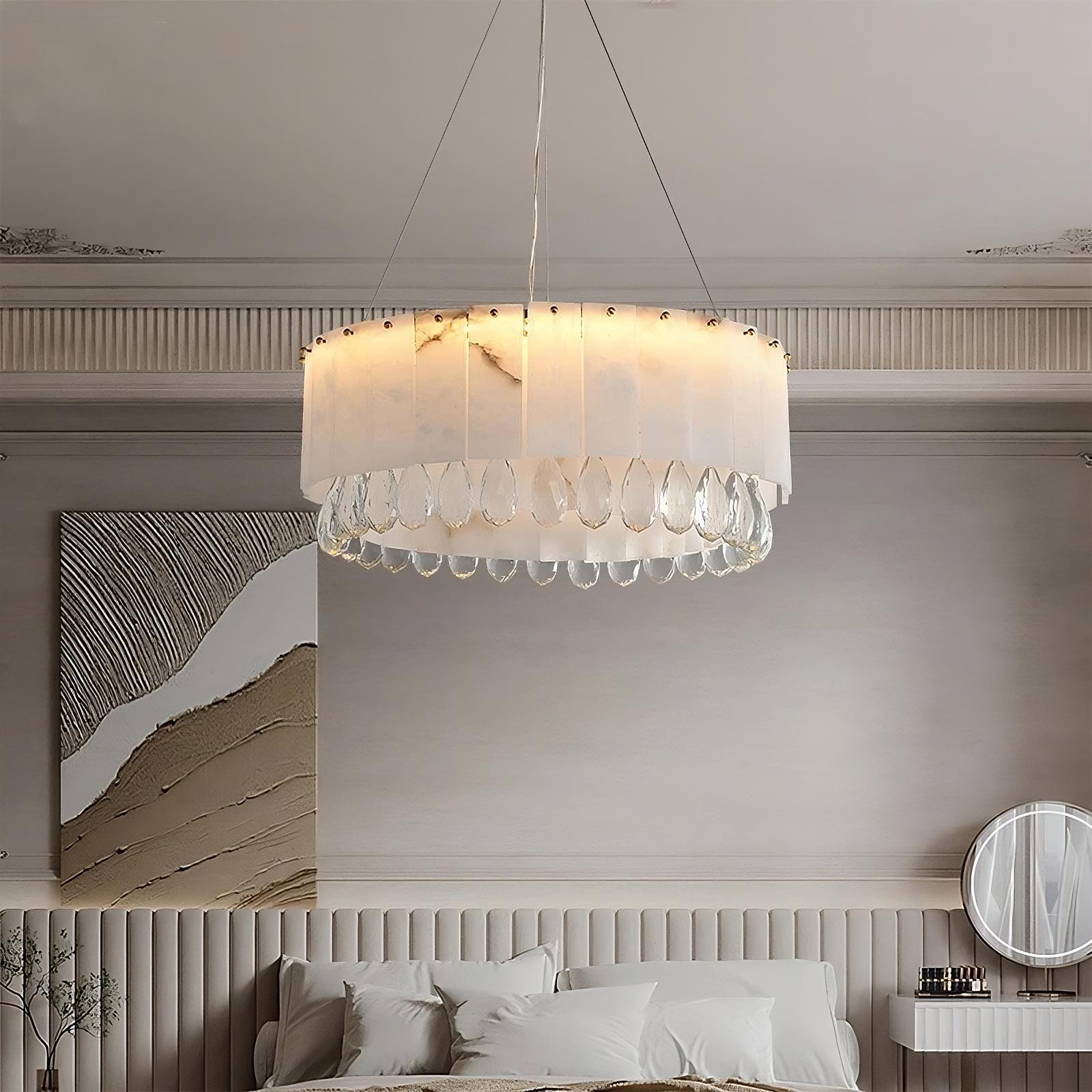 A stylish bedroom features a Modern Crystal and Spanish Marble Chandelier by Morsale.com with hanging crystal ornaments. The room has a neutral color palette with a beige bed, a painting with abstract earth tones above the headboard, and a round mirror on an elegant vanity table in the corner.