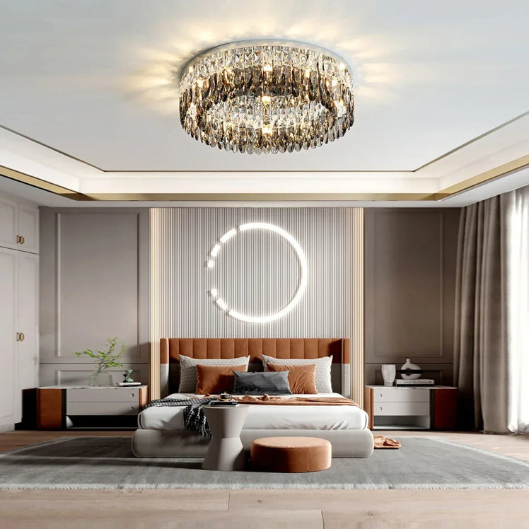 A modern bedroom featuring a large Giano Crystal Ceiling Light from Morsale.com on the ceiling. There is a bed with an upholstered headboard adorned with pillows and a throw blanket. A round illuminated wall decor hangs above the bed. Two nightstands flank the bed, and a large window lets in natural light.
