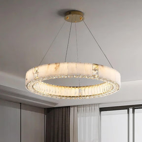 A Morsale.com Natural Marble & Crystal Modern Ceiling Light Fixture with a white and gold finish is suspended from the ceiling by four thin wires. This piece of luxury home décor features translucent natural marble panels, radiating a warm glow. The room has white walls, grey curtains, and large windows.