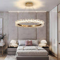 A modern bedroom featuring a Morsale.com Capri Crystal Ceiling Light with crystal accents hanging from the ceiling. The bed is adorned with neutral bedding and two pink accent pillows. Bedside tables with circular lamps are positioned on either side, and a decorative plant is on one table.