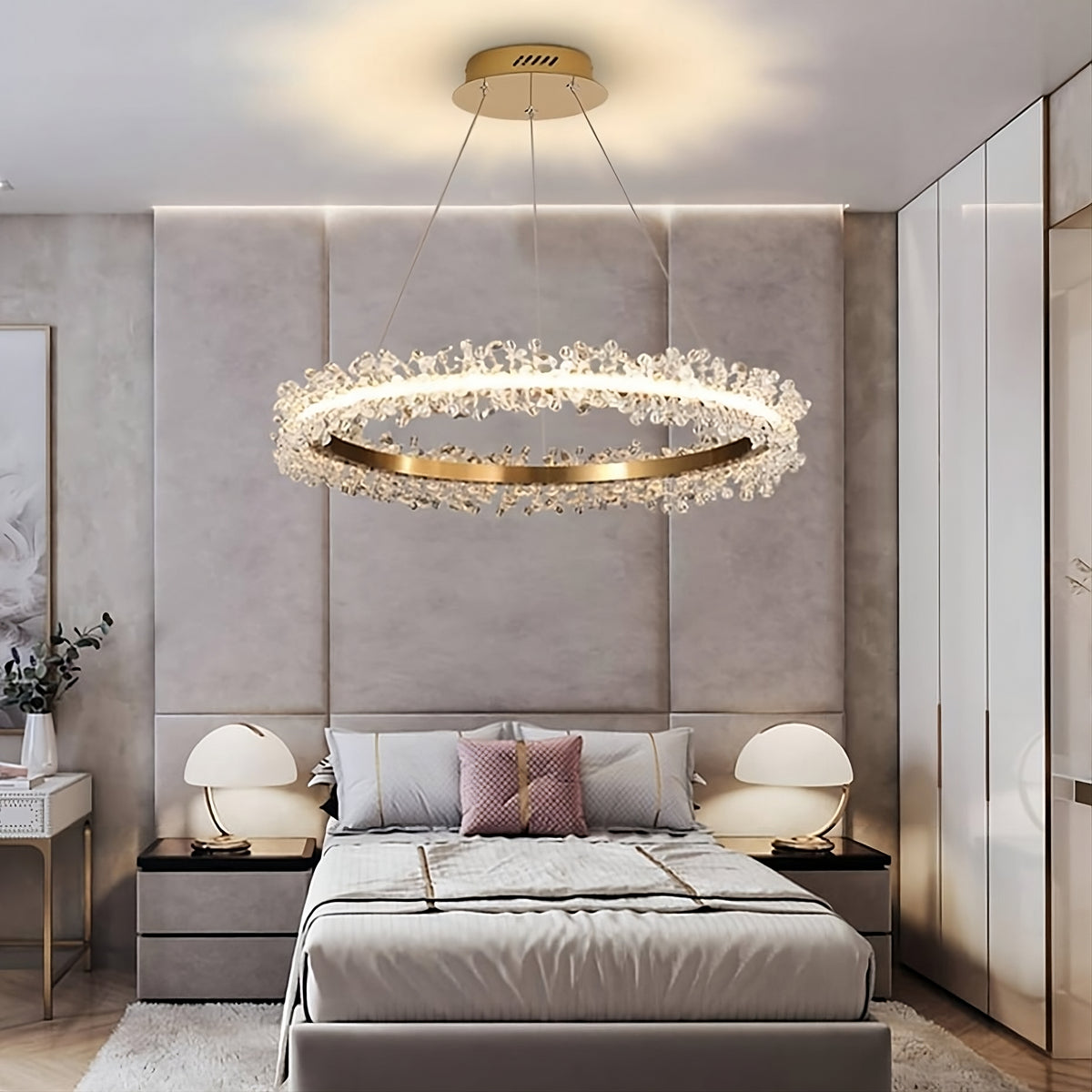 A modern bedroom with a large, circular ceiling chandelier adorned with K9 crystals hanging above a neatly made bed. The **Capri Crystal Ring Chandelier by Morsale.com** flanked by two matching nightstands, each with a lamp featuring a rounded white shade. The room features neutral tones, mirrored closet doors, and a decorative wall behind the bed.