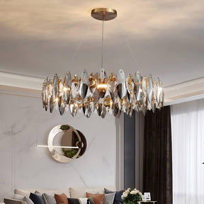 A luxurious living room exudes a modern vibe, featuring the Lazzo Modern Chandelier from Morsale.com, adorned with handmade crystals hanging elegantly from the ceiling. Below, there's a modern circular mirror, a white marble wall, and a comfortable sofa with various pillows. Tall windows allow natural light in.