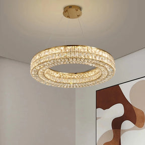 A large, circular Bacci Crystal Modern Chandelier, Gold from Morsale.com hangs from the ceiling, emitting a warm, yellow light. The chandelier has a double-ring design and is gold electroplated. In the background, part of an abstract wall art piece with brown, beige, and white shapes is visible.