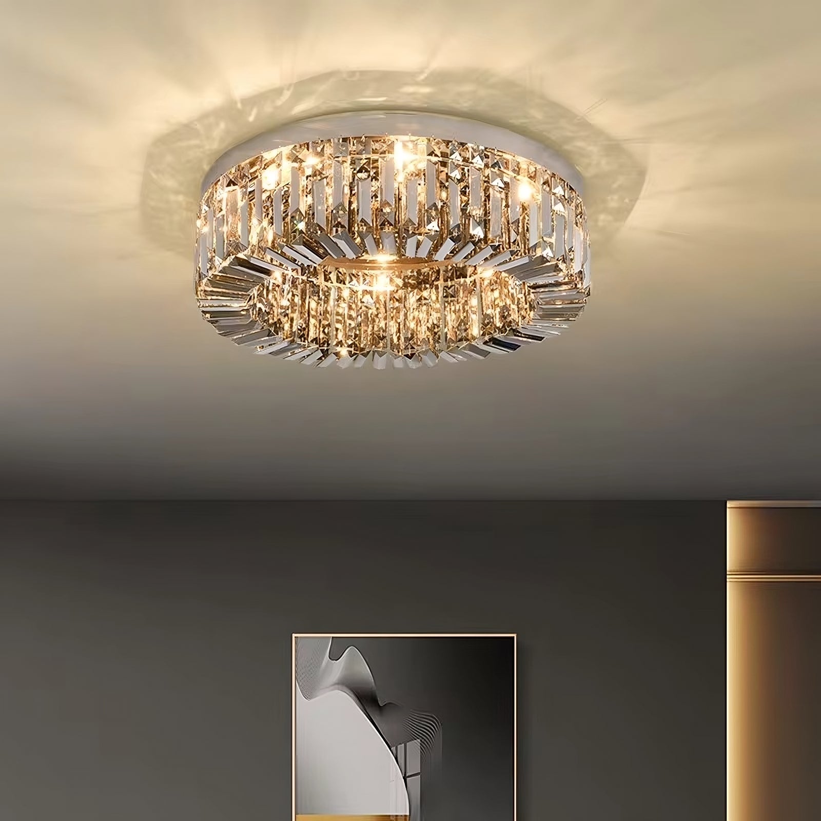 A modern, circular Specchio Smoke Grey Crystal Ceiling Light from Morsale.com is mounted on the ceiling, emitting a warm, soft light. The chandelier features a ring of smoke grey crystals that reflect and disperse the light. Below, a minimalist framed artwork is partially visible on a dark wall, enhancing the modern decor.