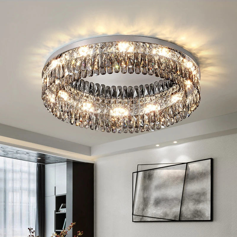 A modern, circular Giano Crystal Ceiling Light from Morsale.com with cascading crystal droplets hangs from the ceiling, emitting a bright light that illuminates the room. The decor includes abstract wall art and a partially visible window with sheer curtains, adding a touch of classic luxury.
