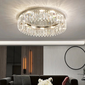 A contemporary living room featuring the Gio Flush Mount Crystal Ceiling Light from Morsale.com on the ceiling with E14 LED bulbs. The space includes modern decor with wood paneling, wall sconces, a striped wallpaper section, and minimal black and white furniture. An open book and plant sit on the coffee table.