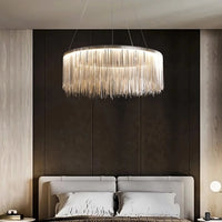 A modern bedroom features the Cadena Modern Chandelier from Morsale.com, a striking circular LED fixture with hanging strands above a gray upholstered bed with neutral bedding, set against a dark accent wall and light wood panels.