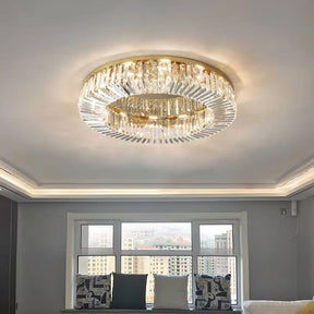 A luxurious modern living room showcases the Specchio Crystal Ceiling Light Fixture from Morsale.com, featuring a large circular chandelier with cascading crystal prisms and a gold frame. The ceiling is subtly lit with recessed lighting, enhancing the modern decor. Below the window seat, a panoramic cityscape view is visible through a wide window.