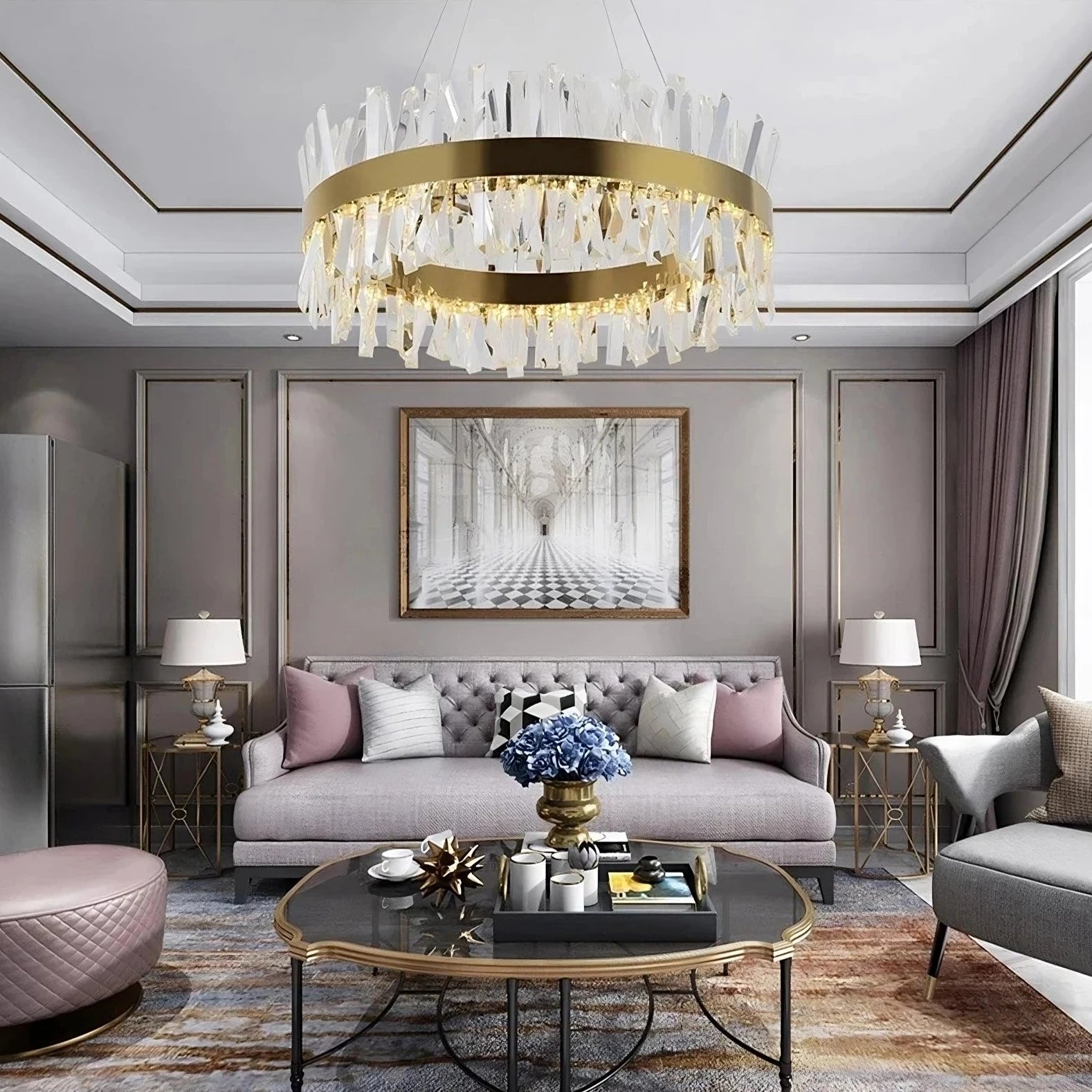 A modern living room with a tufted light gray sofa, decorative pillows, and a glass coffee table adorned with a floral arrangement. A large Calvi Crystal Chandelier from Morsale.com, featuring gold and clear crystal, hangs from the ceiling. Elegant side tables with lamps, framed artwork, and large windows complete the space.