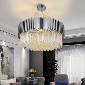 A modern living room features the elegant lighting of a Morsale.com Gio Crystal Contemporary Chandelier, showcasing vertical chrome and crystal rods hanging from a circular frame. The room includes a large abstract painting, plush gray sofas with cushions, and a gold table lamp on a side table. A sheer curtain covers the window.