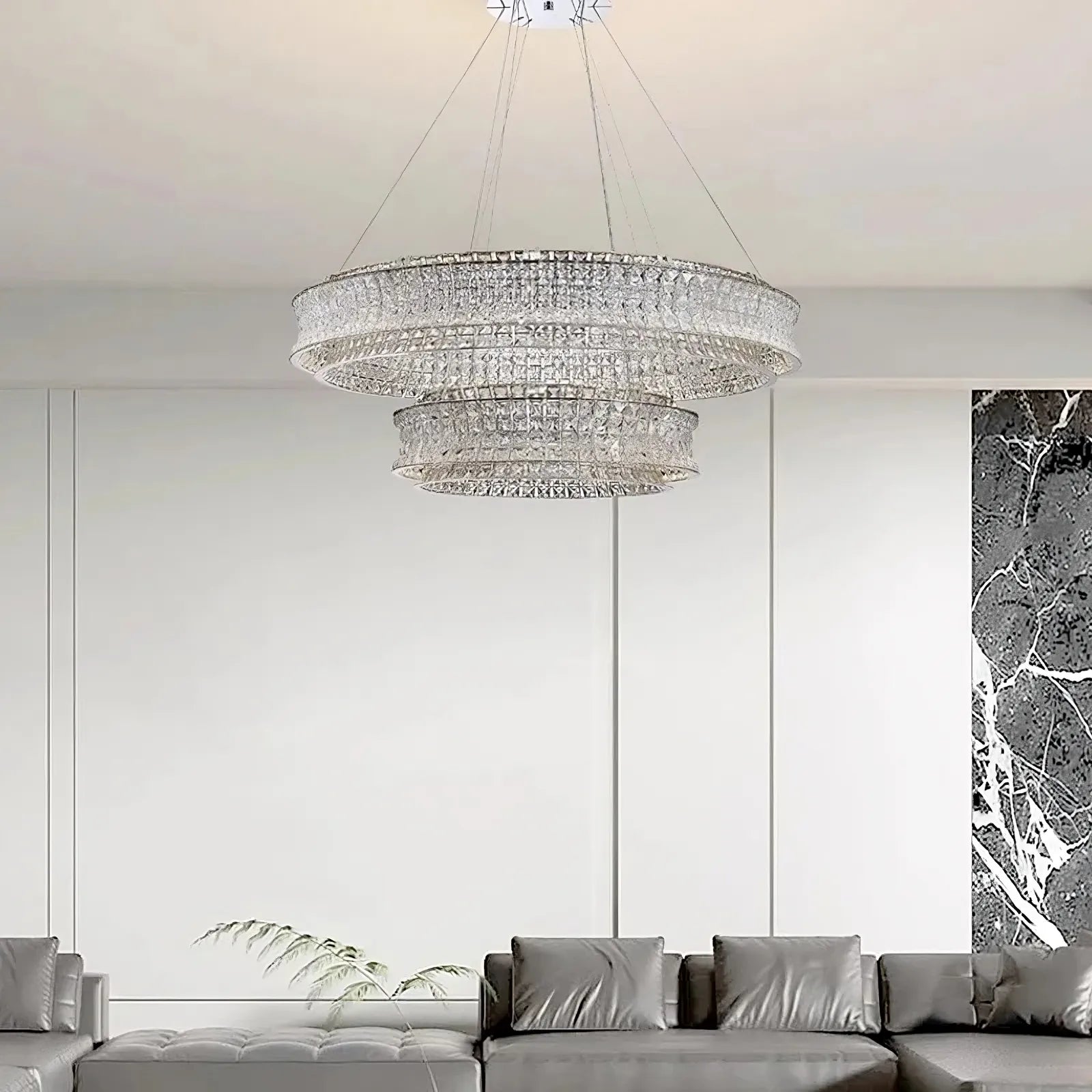 A modern living room features the Morsale Bacci Modern Crystal Chandelier, its sparkle amplified by LED bulbs. Below, a gray sectional sofa teems with pillows. White paneled walls and a black marble accent on the side complete this contemporary space.
