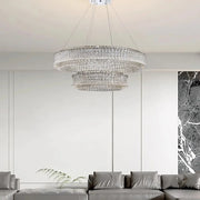 A modern living room features the Morsale Bacci Modern Crystal Chandelier, its sparkle amplified by LED bulbs. Below, a gray sectional sofa teems with pillows. White paneled walls and a black marble accent on the side complete this contemporary space.
