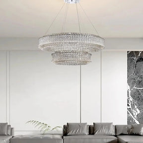 A modern living room features the Morsale Bacci Modern Crystal Chandelier, its sparkle amplified by LED bulbs. Below, a gray sectional sofa teems with pillows. White paneled walls and a black marble accent on the side complete this contemporary space.

