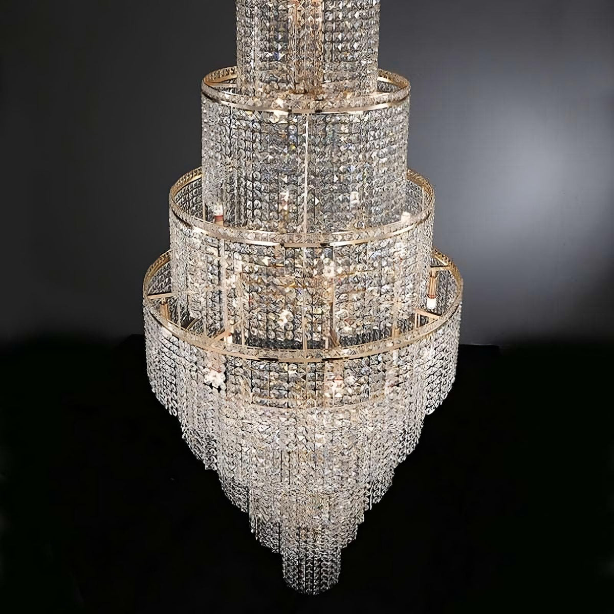 The Alleri Crystal Foyer Chandelier, Gold from Morsale.com is an elaborate multi-tiered lighting masterpiece. It features intricate detailing with cascading strands and multiple circular layers adorned with sparkling crystals and elegant crystal shades, epitomizing modern luxury against a dark background.