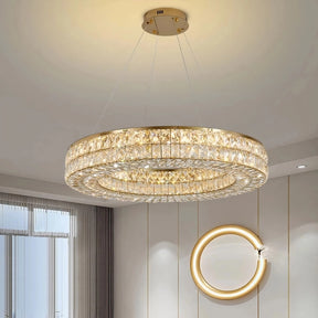 A sophisticated Bacci Crystal Modern Chandelier, Gold by Morsale.com hangs from the ceiling, emitting a warm, golden light. Below it, a polished, gold-framed mirror decorates the wall, reflecting light into the elegantly furnished room, which features large windows with sheer curtains.