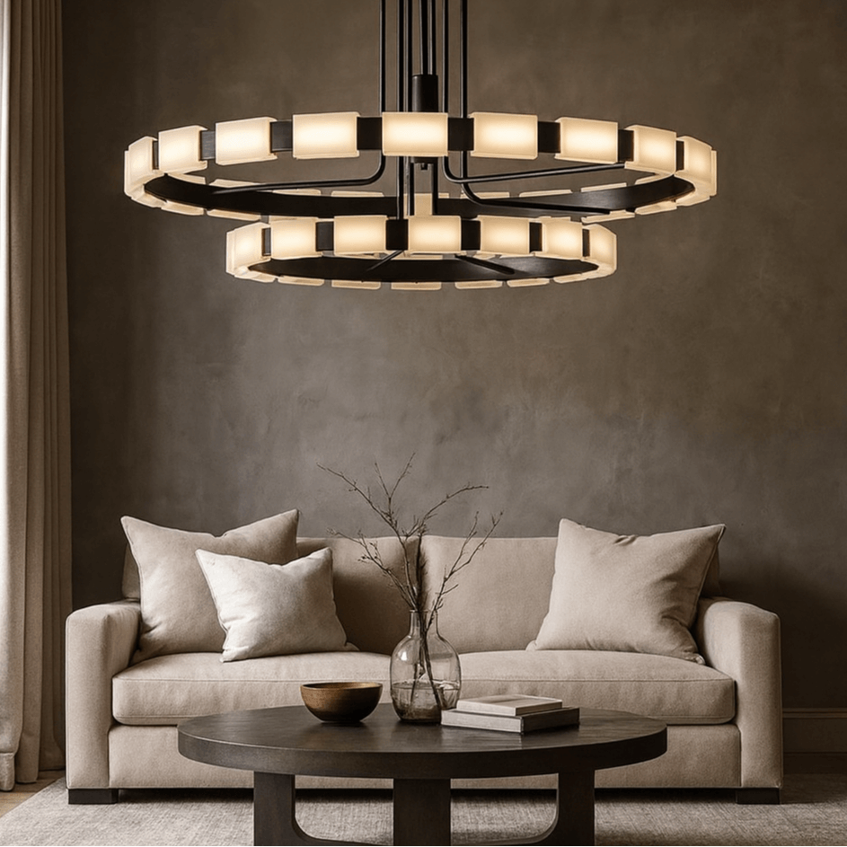 A modern living room features a beige sofa, neutral pillows, a round dark coffee table with minimalist decor, and the Aveline Modern Chandelier by Morsale.com glowing above against a taupe textured wall.