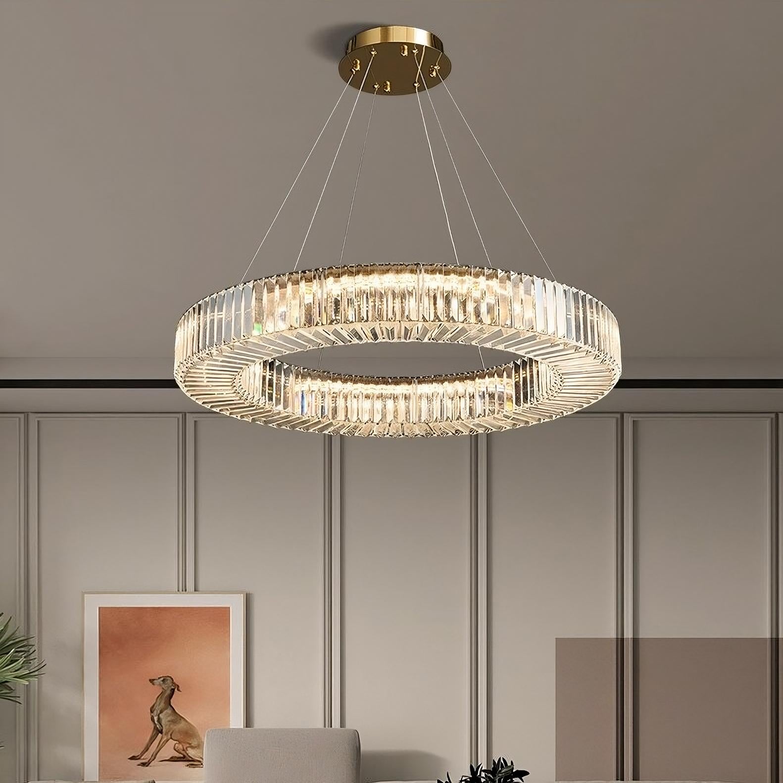 An Azali Crystal Ceiling Light from Morsale.com, featuring a modern circular design with a gold rim and clear crystals, hangs elegantly from the ceiling, supported by thin wires. Below the chandelier is a framed picture of a dog, a plant, and a wall adorned with minimalist panel designs in a contemporary room.