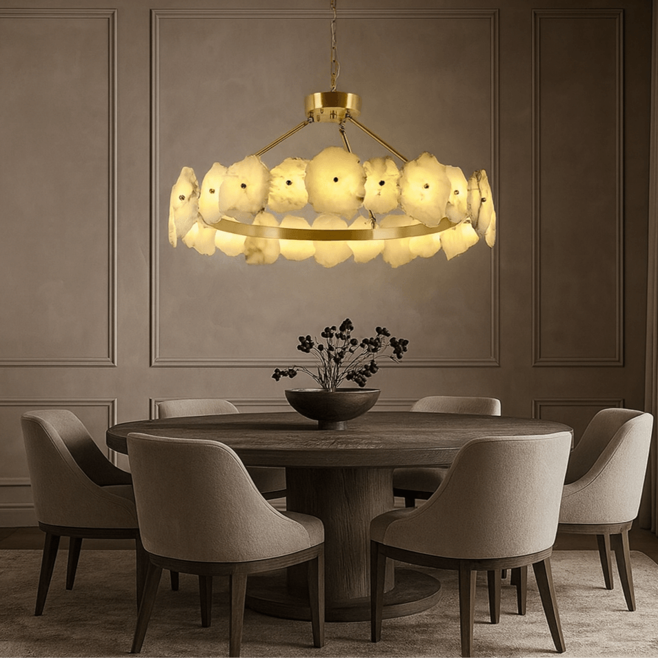 A round wooden dining table with six upholstered chairs sits beneath the Bigman Corton Marble & Brass Chandelier in a softly lit, neutral-toned room with paneled walls.