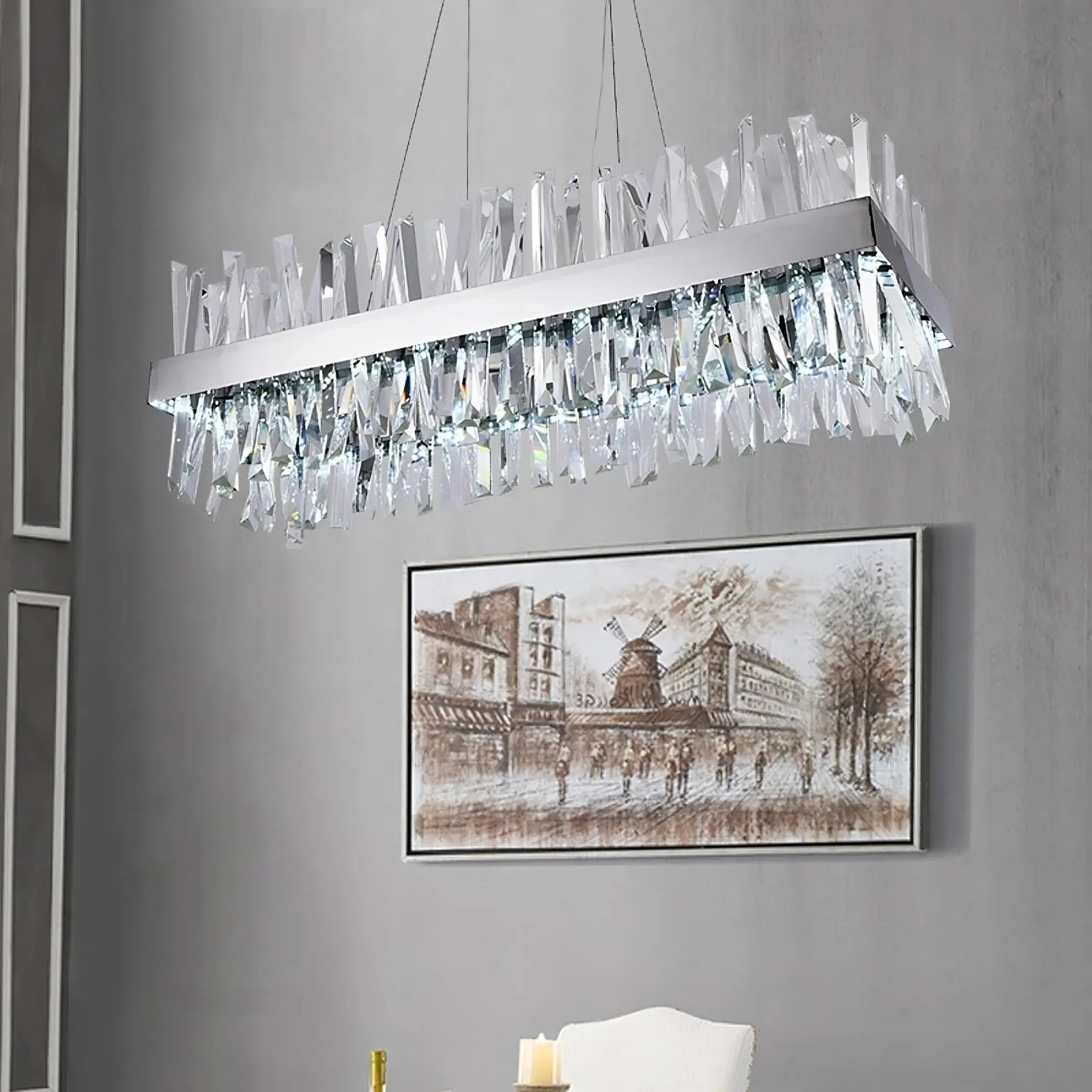 A modern rectangular Calvi Crystal Dining Room Light Fixture from Morsale.com, adorned with unique, jagged K9 crystal pieces, hangs from the ceiling in a contemporary room. Below it is a white chair and a candle. On the wall, there’s an art piece depicting an old street scene with buildings and people walking.