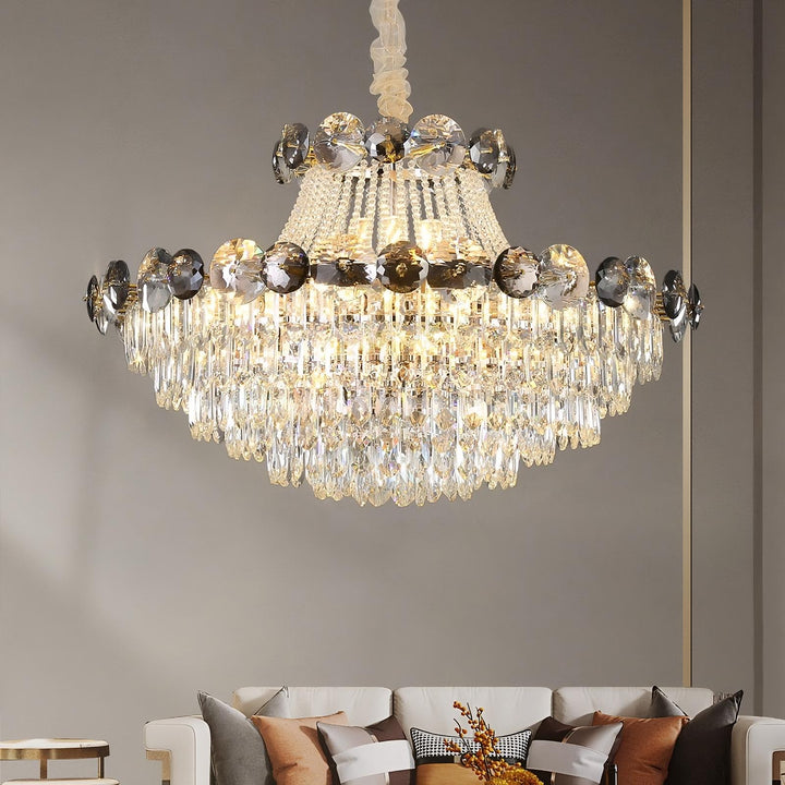 A luxurious Morsale Gioro Contemporary Chandelier with dimmable LED lighting hangs from the ceiling, casting a warm glow. It features rows of dangling crystals and circular reflective elements. Below, a stylish living room is partially visible, showcasing a sofa with decorative pillows and a coffee table.