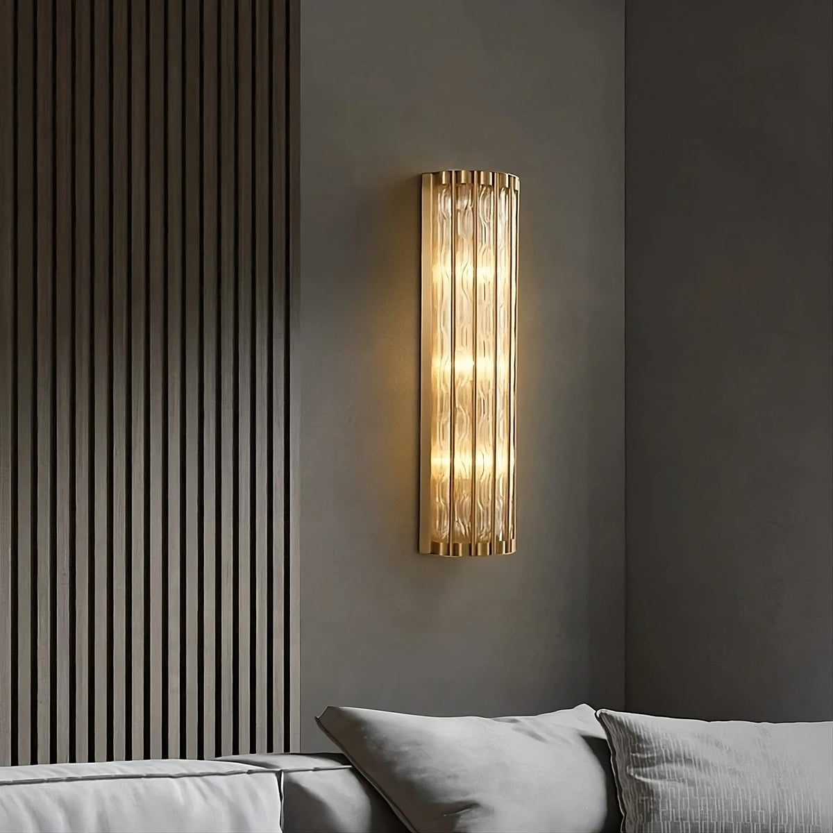 A modern, vertically oriented Modern Copper & Glass Wall Sconce by Morsale.com with a ribbed pattern and warm yellow LED bulbs is mounted on a dark gray wall beside a sofa with light gray cushions. The energy-efficient illumination adds a cozy ambiance to the minimalist room.