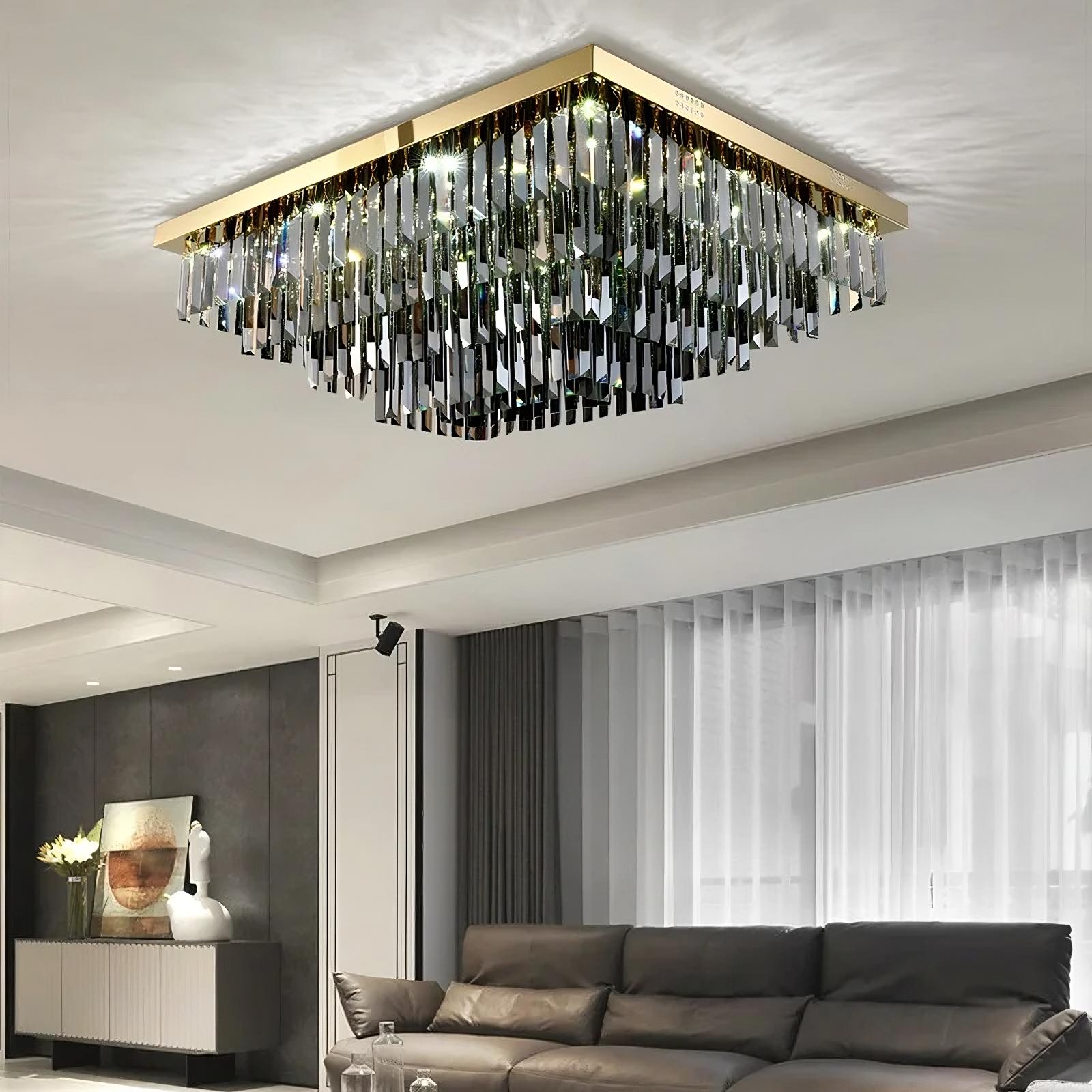 A modern living room featuring a large, rectangular Gio Smoke Grey Crystal Ceiling Chandelier by Morsale.com hanging from the ceiling. The chandelier has multiple dangling pieces, reflecting light. Below it, a comfortable brown leather sofa is positioned near large windows with sheer curtains, showcasing an elegant design.