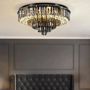 A luxurious Gio Crystal Ceiling Light from Morsale.com, featuring cascading, elongated glass prisms and smoke grey crystals, hangs from the ceiling in a room with gray walls and a tufted gray headboard. The elegant lighting fixture adds a touch of sophistication and modern design to the space.