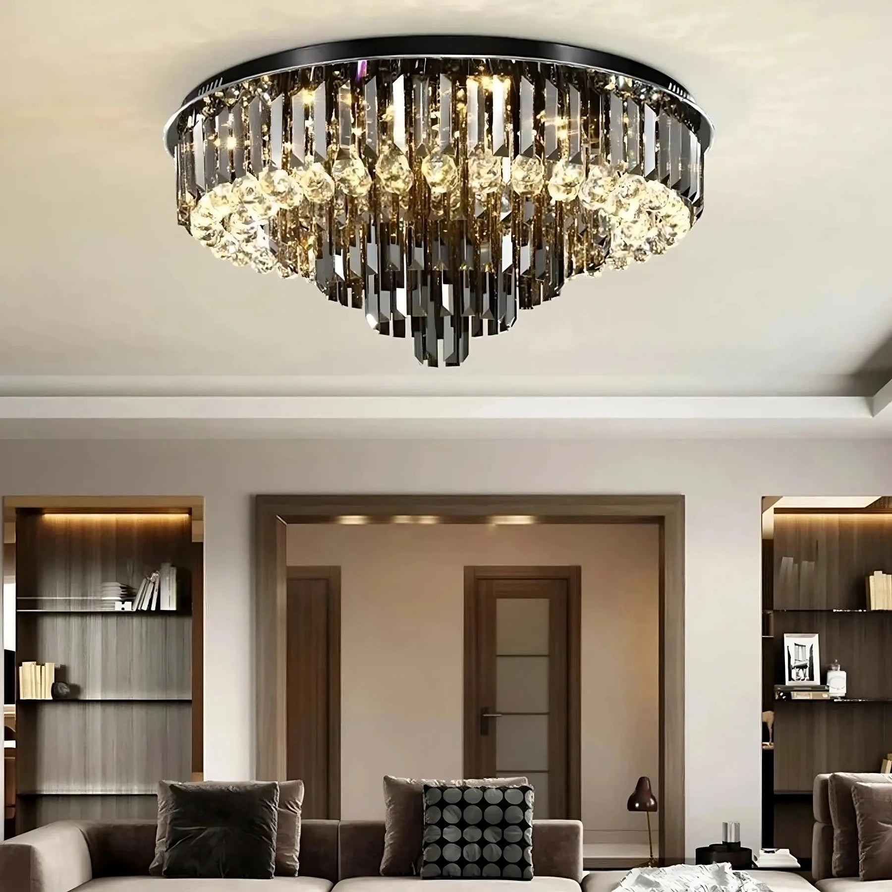 A modern living room features a large Gio Crystal Ceiling Light from Morsale.com, adorned with smoke grey crystals and stainless steel accents, hanging prominently from the center of the ceiling. The room boasts a contemporary design with built-in shelving units, neutral-colored furniture, and open doorways leading to adjacent spaces.