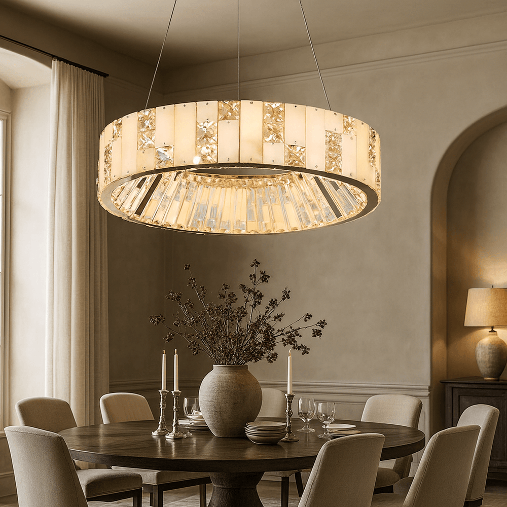 A round table with beige chairs sits beneath the Morsale Edes Marble & Crystal Chandelier, featuring gold and glass details, in an elegant dining room with neutral walls and a vase of branches as the centerpiece.