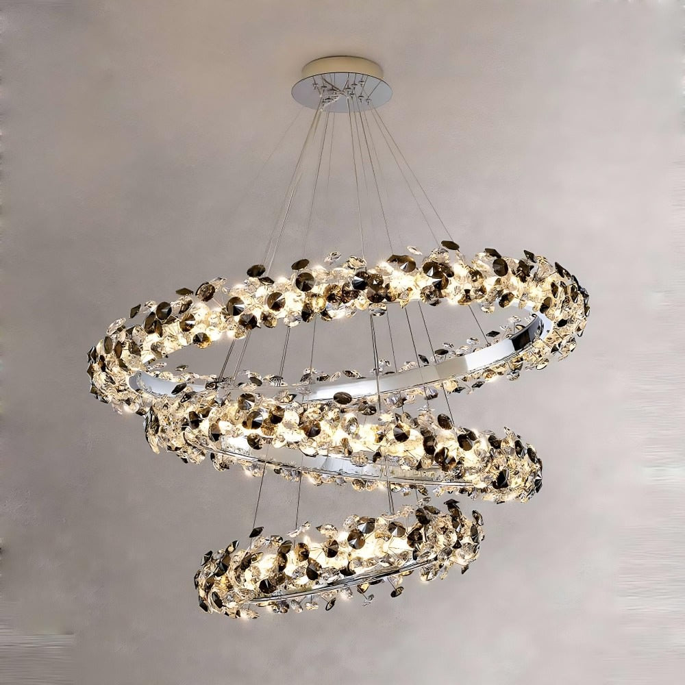 The Di Gardo Crystal Modern Chandelier by Bigman is a sophisticated lighting fixture featuring three cascading, circular rings adorned with numerous sparkling crystals and small metal leaves. The rings are suspended from the ceiling with thin wires and showcase an LED light source that creates an elegant, floating effect against a neutral background.