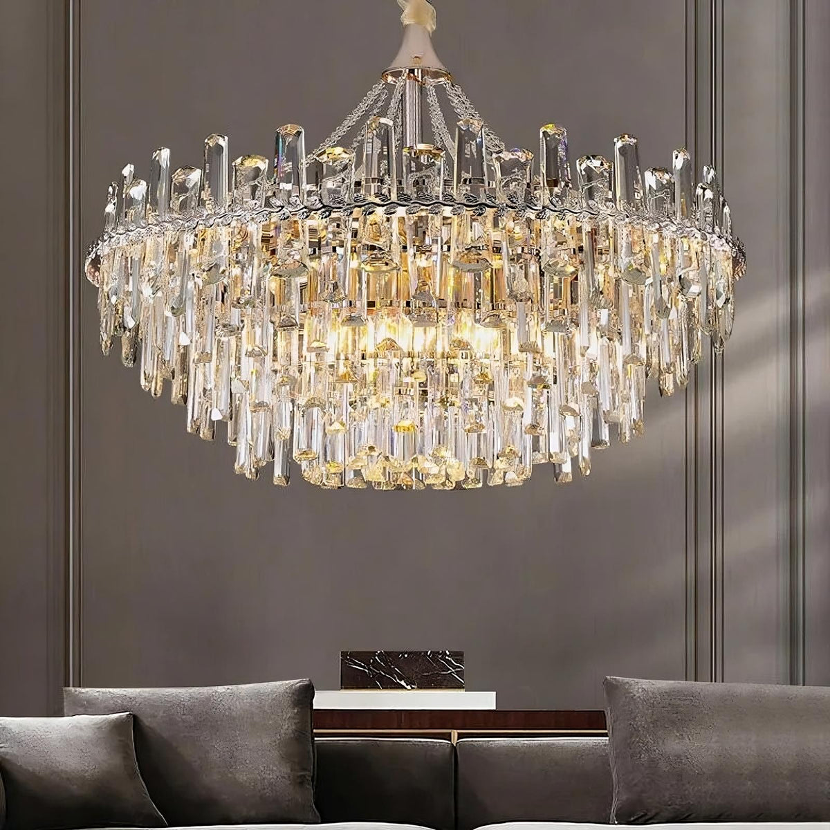 An elegant, contemporary Gio Crystal Modern Chandelier by Morsale.com hangs from the ceiling, featuring numerous handmade crystals that refract light beautifully. Below, a modern sofa with plush gray cushions contrasts against a backdrop of subtly paneled grey walls.
