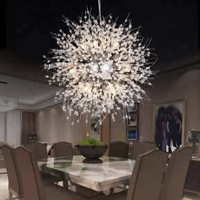 A dining room with a rectangular table surrounded by eight chairs. The centerpiece is an El Sol Crystal Dandelion Lighting Fixture by Morsale.com, featuring intricate K9 crystals and G9 LED bulbs that emit a warm light. A decorative bowl filled with green apples sits on the marble table, while a group photo hangs on the wall.